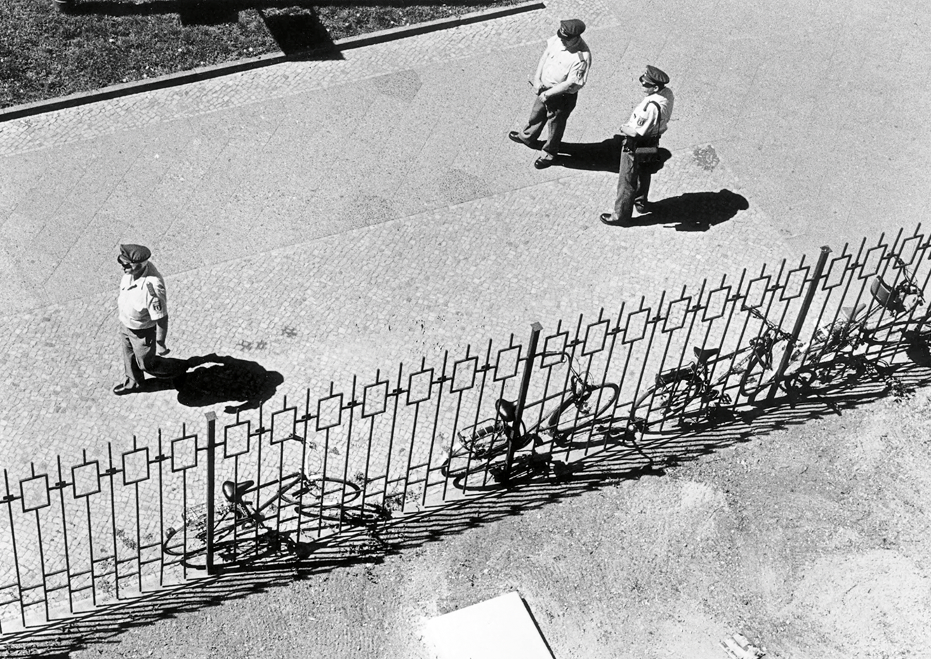 Wachpersonal der Berliner Polizei vor der kubanischen Botschaft, welche sich bis 1991 in der Berliner Straße befand. - Waisenhaus Pankow Drei Personen in Uniform gehen entlang einer gepflasterten Straße, während Fahrräder hinter einem Zaun stehen.