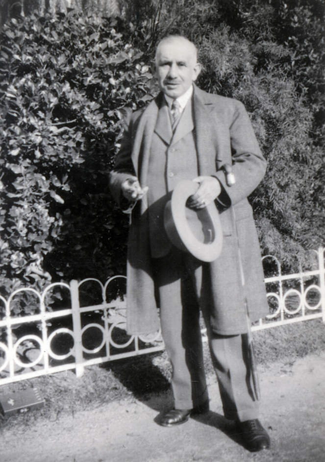 Baumeister Alexander Beer - Waisenhaus Pankow Ein älterer Mann in Anzug und Hut steht vor einer Hecke, hält einen Stock in der Hand.