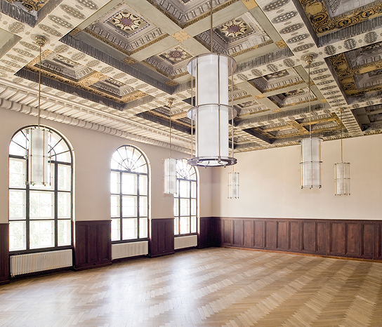 Wandpaneele wurden wieder angebracht, welche auch ursprünglich den Betsaal zierten. - Waisenhaus Pankow Heller, leerer Raum mit Holzfußboden, großen Fenstern und kunstvollem Deckenmuster, beleuchtet von modernen Hängeleuchten.