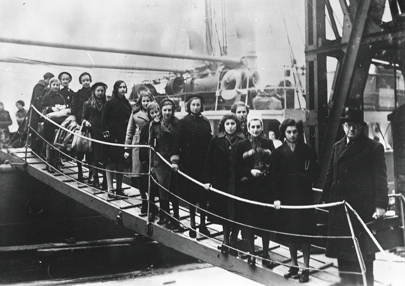 Kindertransporte nach Großbritannien: Jüdische Kinder bei ihrer Ankunft mit der „Warschau“ in London im Februar 1939. - Waisenhaus Pankow Gruppierung von Mädchen, die auf einer Brücke zu einem Schiff gehen, umgeben von historischen Elementen.