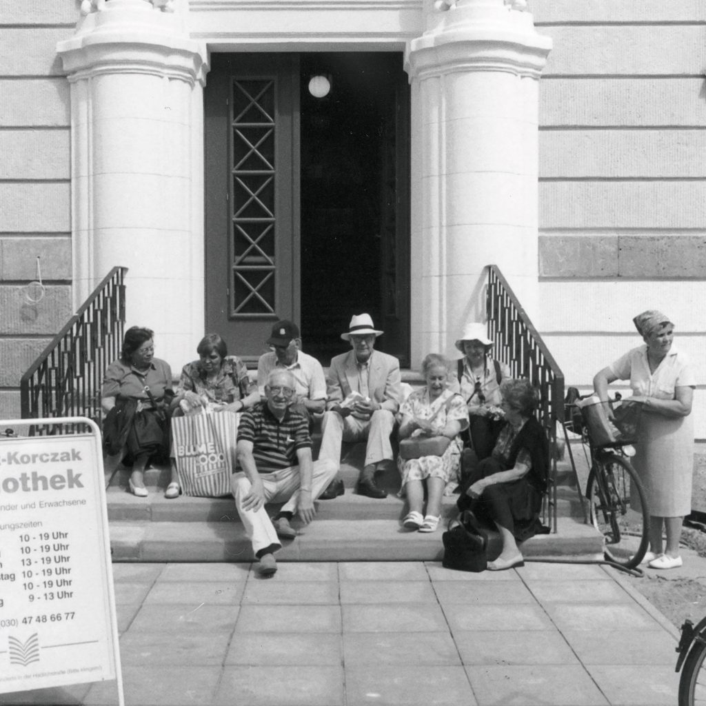 Begegnung am Waisenhaus - Waisenhaus Pankow Gruppierung von Menschen auf den Stufen eines Waisenhauses, einige sitzen, andere stehen, mit einem Schild der Apotheke im...