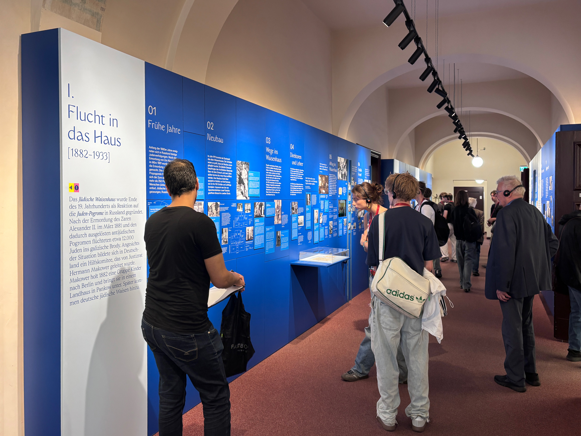 Ausstellungsansicht 1 - Waisenhaus Pankow Besucher betrachten eine Ausstellung mit blauen Wänden, die Informationen und Bilder zu Flucht und Asyl zeigen.