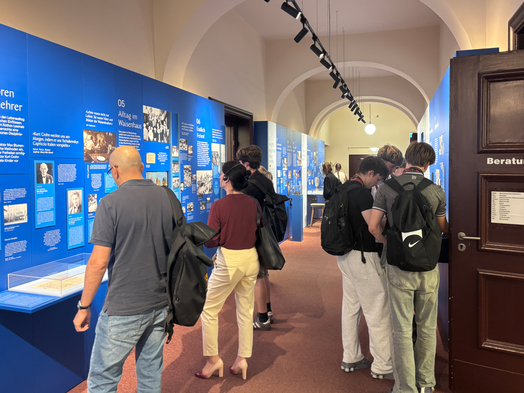 Ausstellungsansicht 4 - Waisenhaus Pankow Besucher betrachten eine Ausstellung mit blauen Wänden und Informationsständen in einem langen Flur.